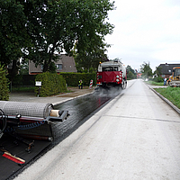 Een termmachine rijdt vooruit, gevolgd door het aanbrengen van SamiGrid® asfaltwapening op de weg