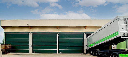 Lubratec Stabidoor Drie gesloten groene Lubratec Stabitore bij een beige magazijn. Een vrachtwagen wordt uitgeladen bij de open poort rechts.